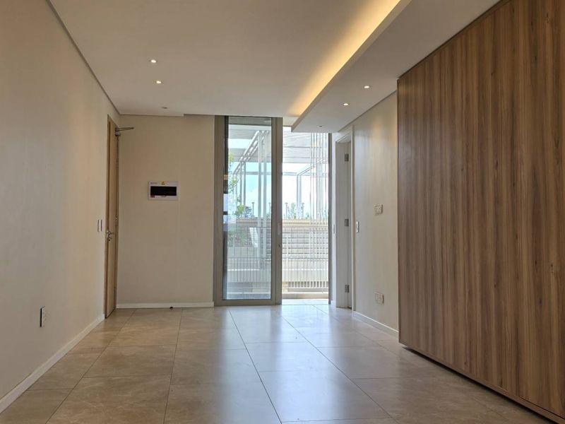 A modern, well-lit hallway featuring tiled flooring, recessed ceiling lights, and sleek built-in wood-finish cabinetry leading to a glass door.