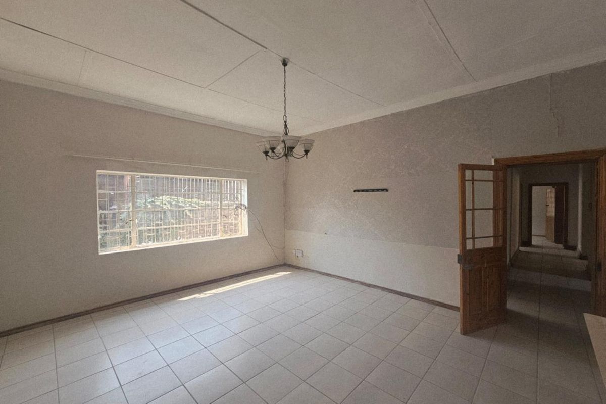An empty living room featuring tiled flooring, a central chandelier, and a large window with security bars, leading into a hallway.