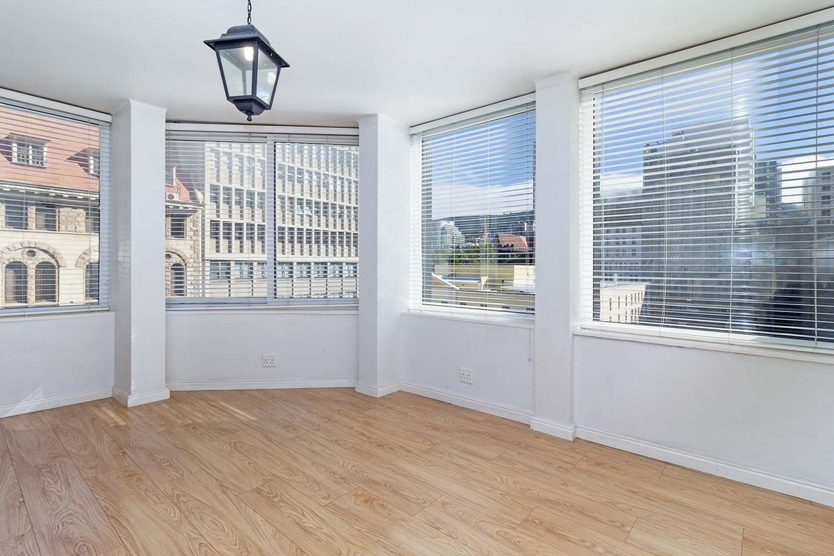 A bright, empty living area featuring light wood-look flooring, large windows with fitted blinds, and an urban city view.