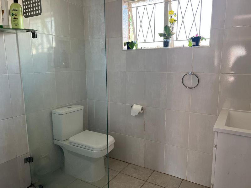 A functional bathroom featuring tiled walls and flooring, a glass shower screen, a toilet, and a vanity basin with a window providing natural light.