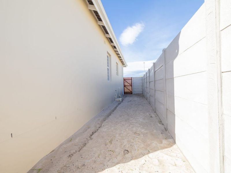 A narrow side passage between the house exterior and a precast concrete boundary wall, featuring a timber gate and sandy ground surface.
