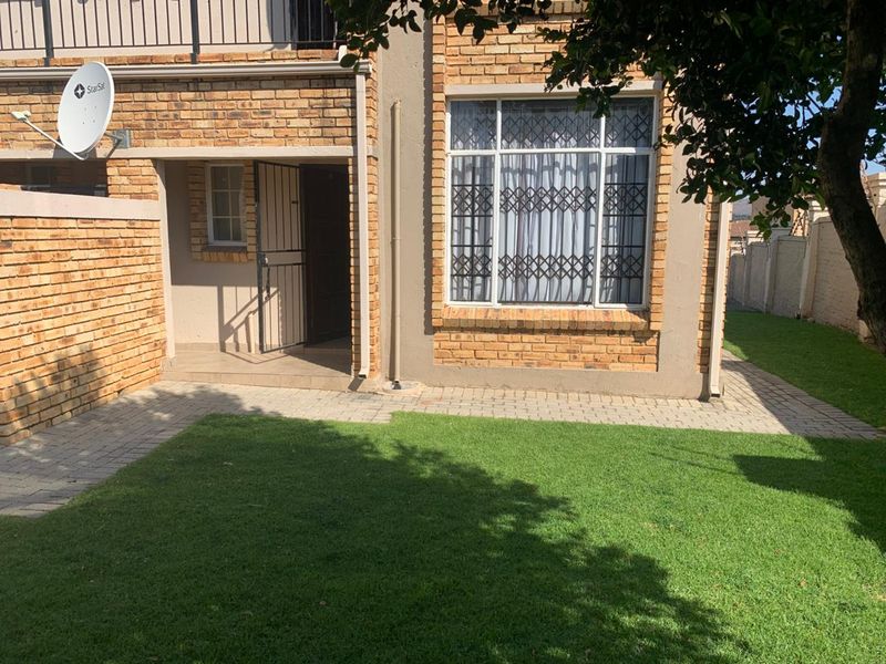 Rear view of the apartment featuring a face brick facade, paved walkway, small lawn area, and secure entrance with security gate and window bars.