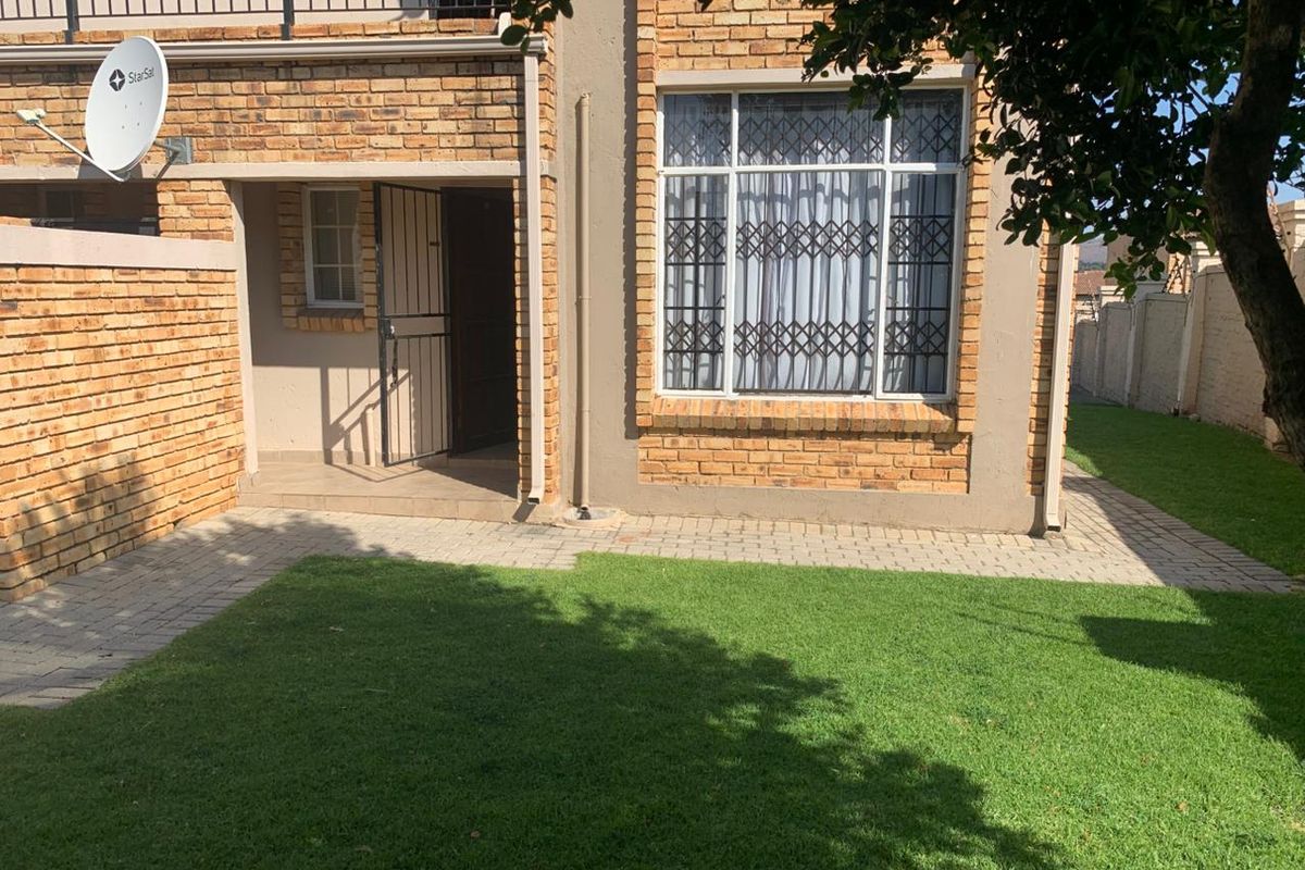 Rear view of the apartment featuring a face brick facade, paved walkway, small lawn area, and secure entrance with security gate and window bars.