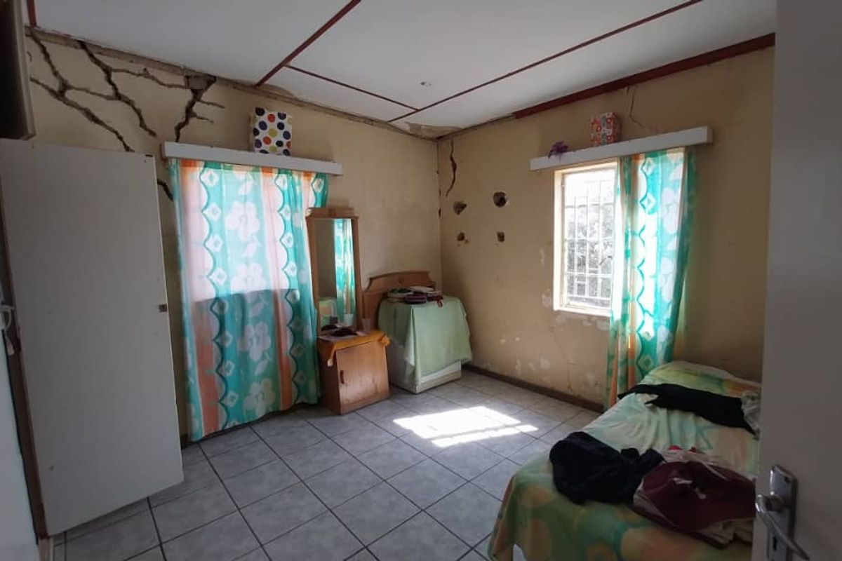 A bedroom featuring tiled flooring and windows with security bars, currently showing significant signs of structural wall cracking and general wear requiring maintenance.
