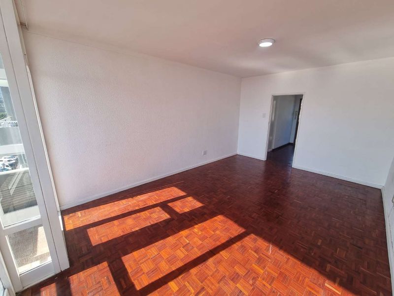 A bright, empty living room featuring classic parquet flooring and a large sliding glass door providing natural light.
