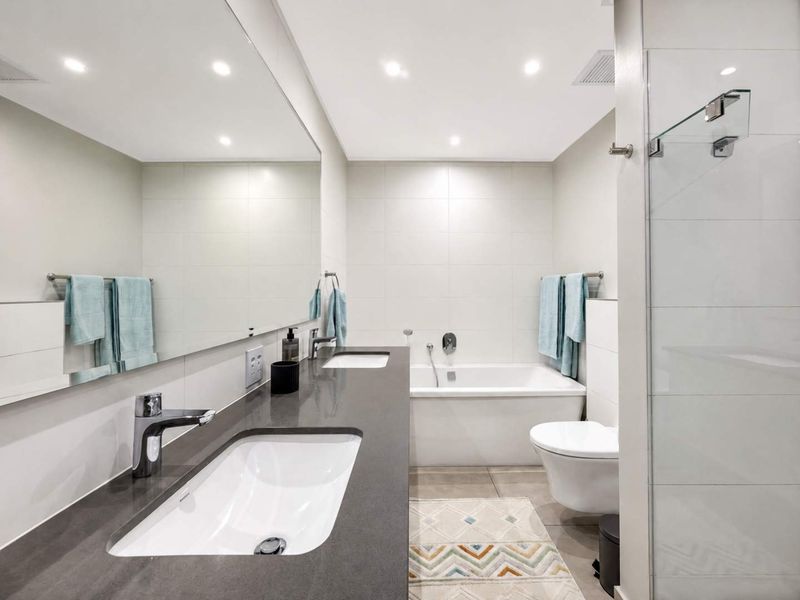 Modern bathroom featuring a double vanity with stone countertop, large wall mirror, built-in bathtub, wall-mounted toilet, and a glass-enclosed shower area.