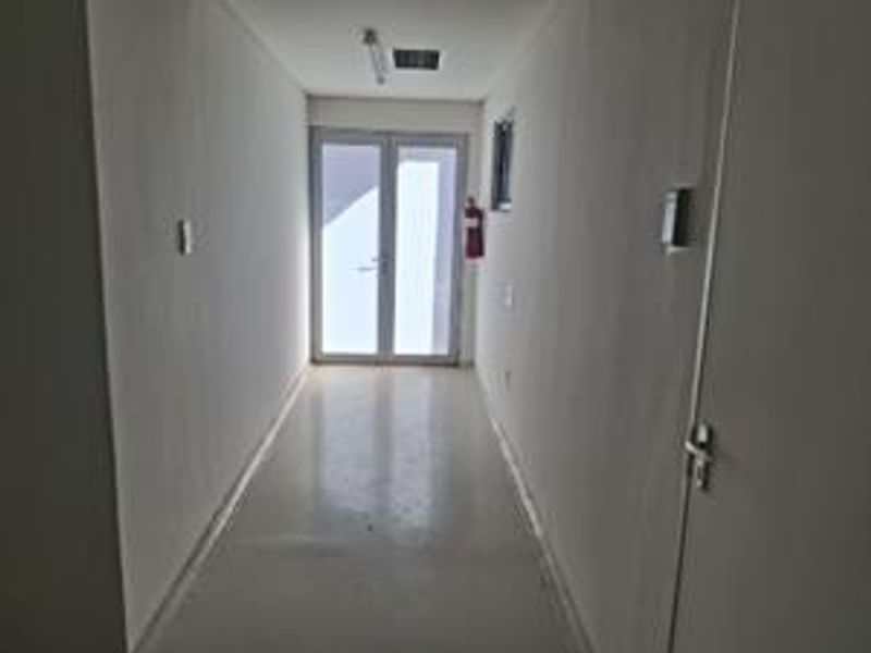 A clean, empty office hallway featuring white walls, smooth flooring, and double glass doors providing natural light access.