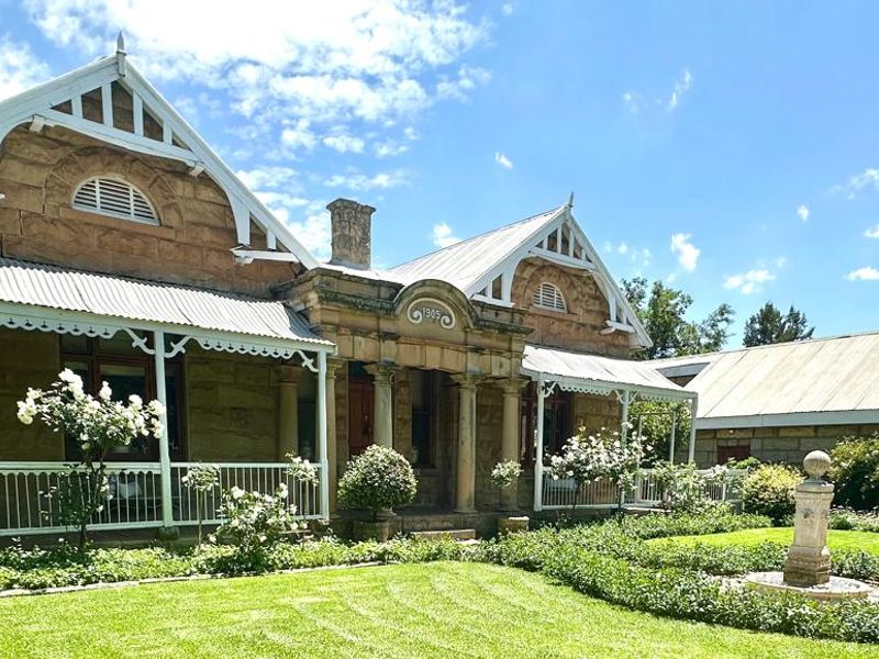 A charming historic stone house featuring a classic gabled facade, a wide covered veranda, and a beautifully landscaped front garden with a stone fountain.