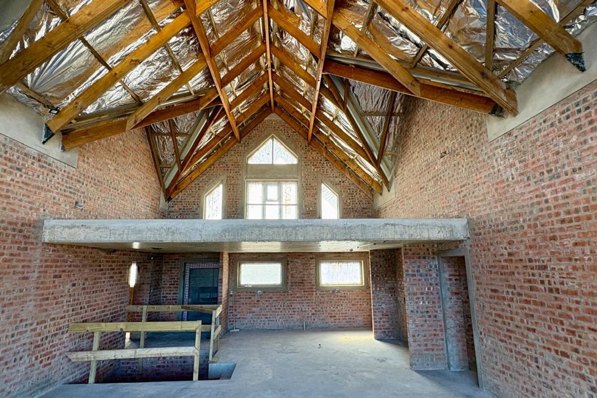 Spacious double-volume interior space under construction, featuring exposed brick walls, timber roof trusses, and a concrete mezzanine level with large feature windows.