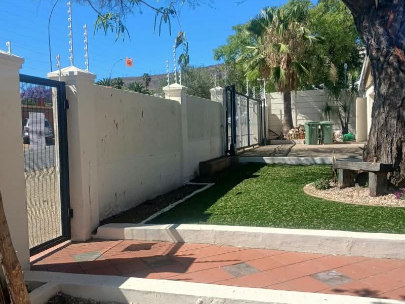 Front exterior area featuring a paved walkway, artificial lawn, perimeter wall with electric fencing, and mature tree.