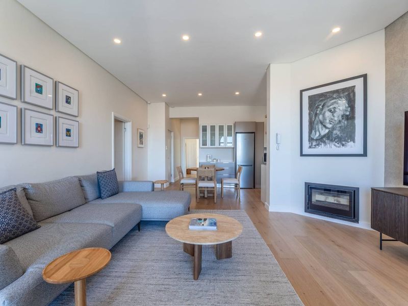 A bright and modern open-plan living area featuring hardwood floors, recessed lighting, and a sleek built-in fireplace, seamlessly connecting to a compact kitchen space.