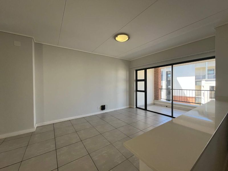 An empty open-plan living area featuring neutral tiled flooring, a breakfast bar, and a large sliding glass door leading to a balcony.