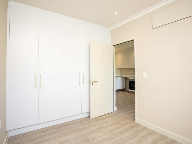 A bright, empty bedroom featuring modern built-in cupboards, light laminate flooring, and a view into the adjacent kitchen area.
