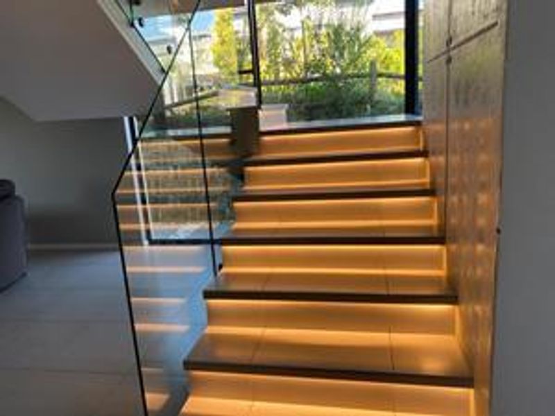Modern staircase featuring a glass balustrade, integrated LED step lighting, and sleek tiled treads, leading to a bright landing area.