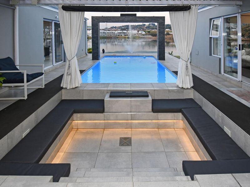 A luxurious sunken outdoor seating area featuring integrated lighting, adjacent to a pristine swimming pool with a water feature and scenic lake views.