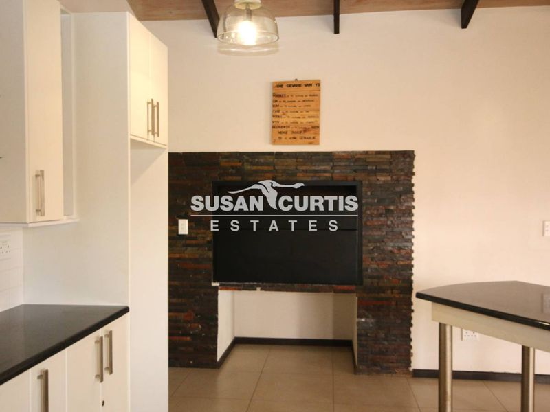 A kitchen area featuring white cabinetry, stone countertops, and a prominent stone-clad fireplace, complemented by exposed ceiling beams and modern pendant lighting.