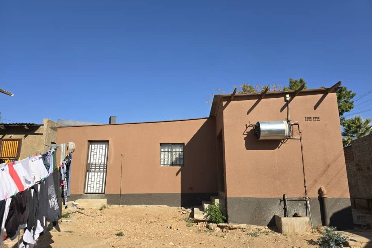 Rear exterior view of the property featuring a brown rendered facade, external geyser unit, security gate, and window with burglar bars.