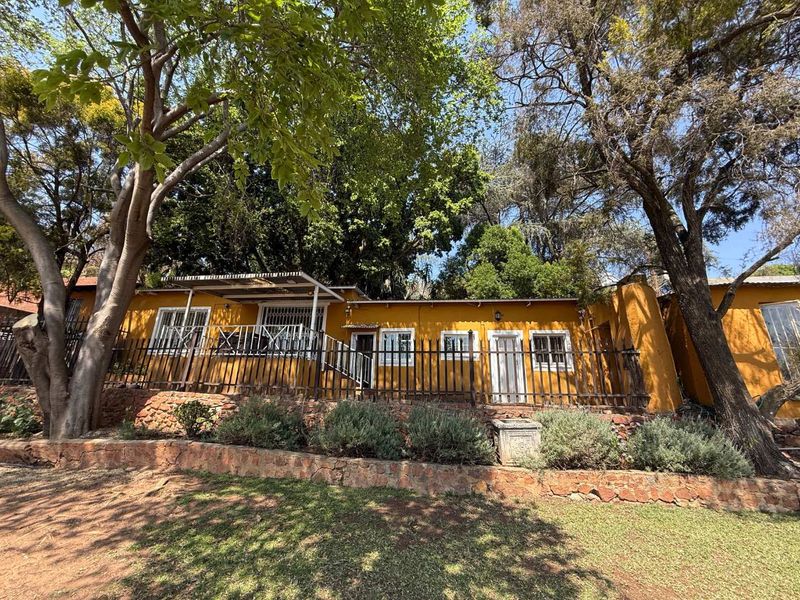 Rear exterior view of the cottage featuring a stone retaining wall, metal security fencing, and a covered patio area surrounded by mature trees.