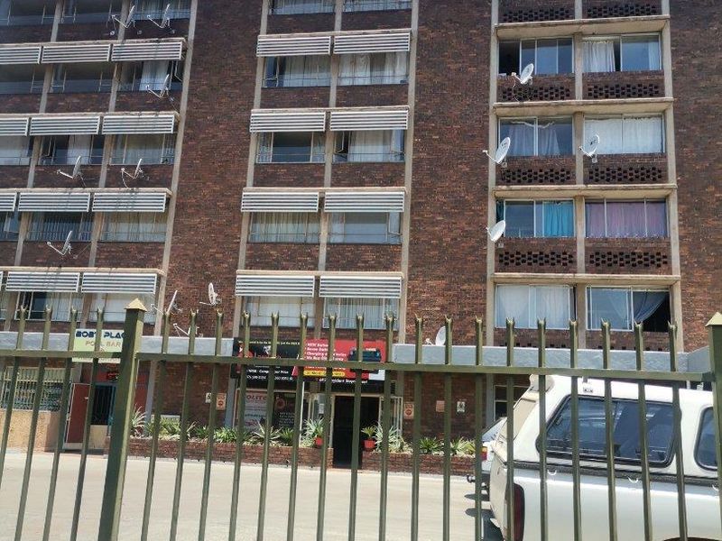 Exterior view of a multi-story brick apartment building featuring metal security fencing, paved driveway, and multiple external satellite dishes.
