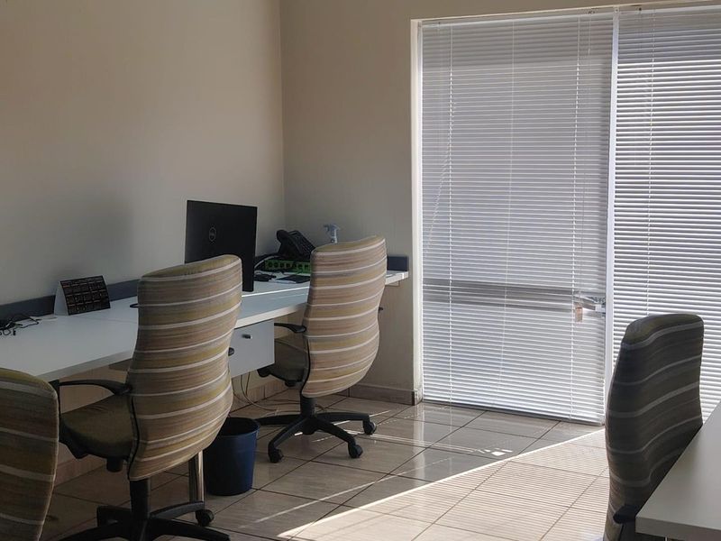 A functional office space featuring a built-in desk, tiled flooring, and large windows fitted with venetian blinds for adjustable natural light.
