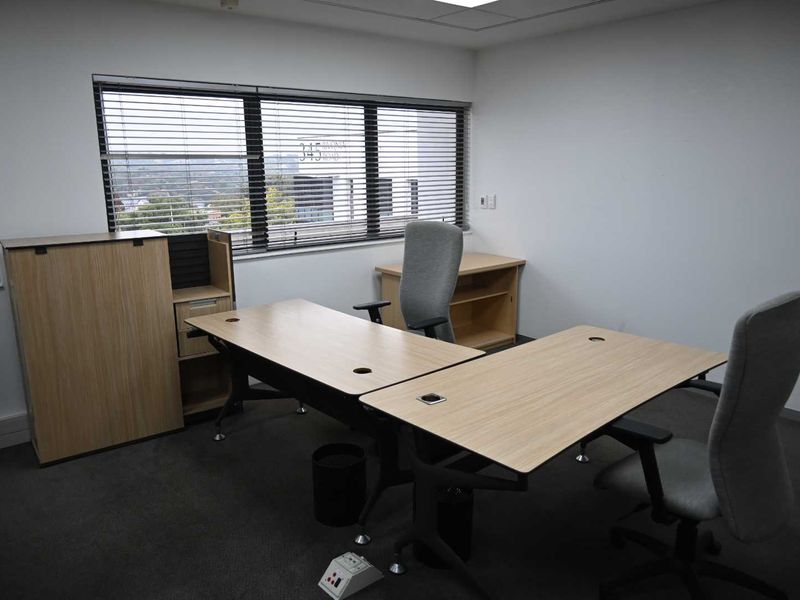 A functional office space featuring carpeted flooring, recessed ceiling lighting, and large windows with venetian blinds providing natural light.