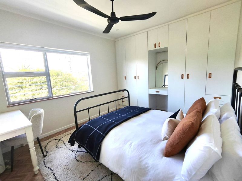 A bright bedroom featuring extensive built-in cabinetry with an integrated vanity nook, a ceiling fan, and large windows providing natural light.