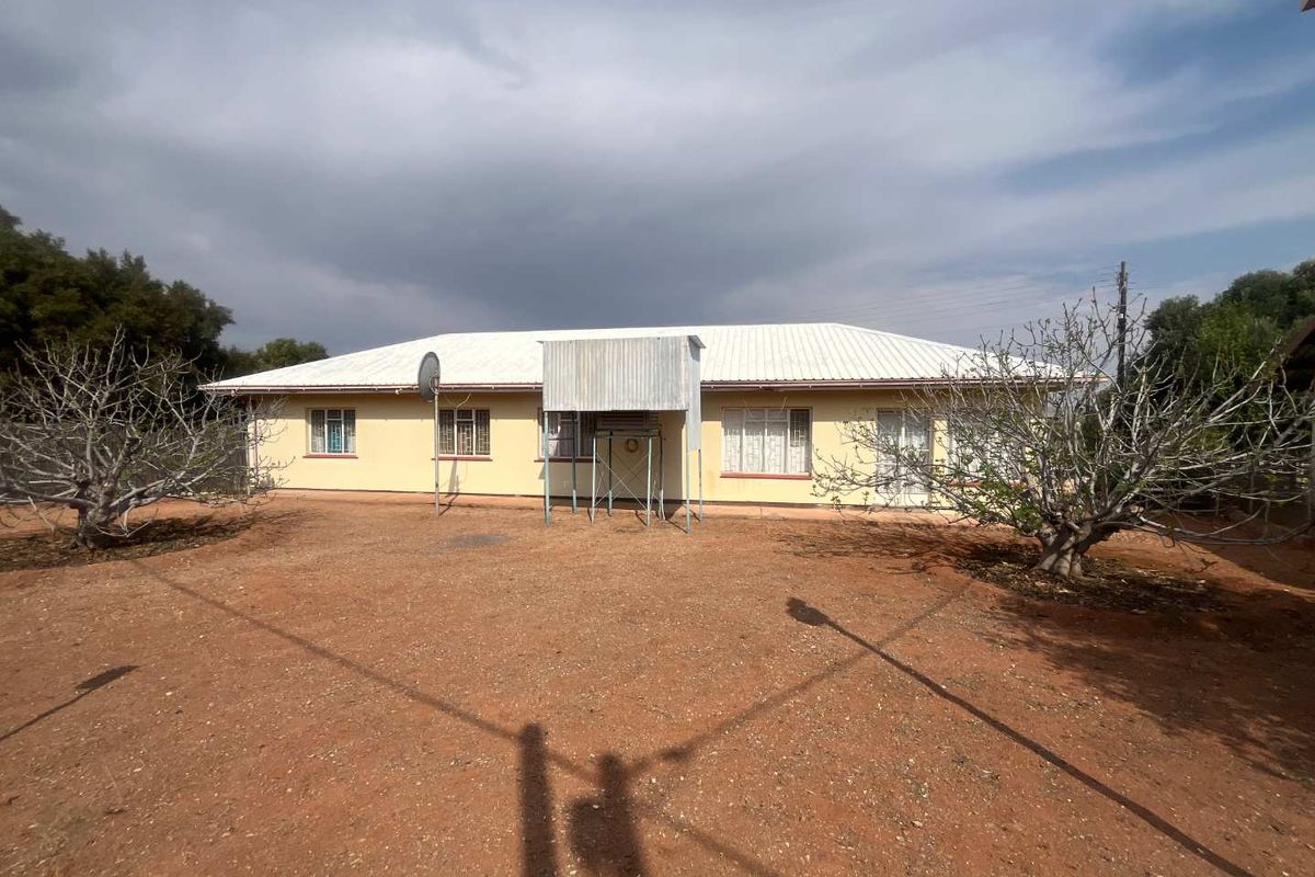 Rear view of a single-story house featuring a corrugated metal roof, security bars on windows, and a metal entrance canopy over a large dirt yard.