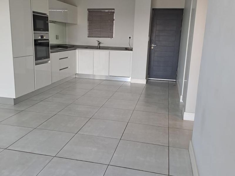 A modern, empty kitchen featuring white high-gloss cabinetry, built-in appliances, grey tiled flooring, and neutral walls, offering a clean and functional space.