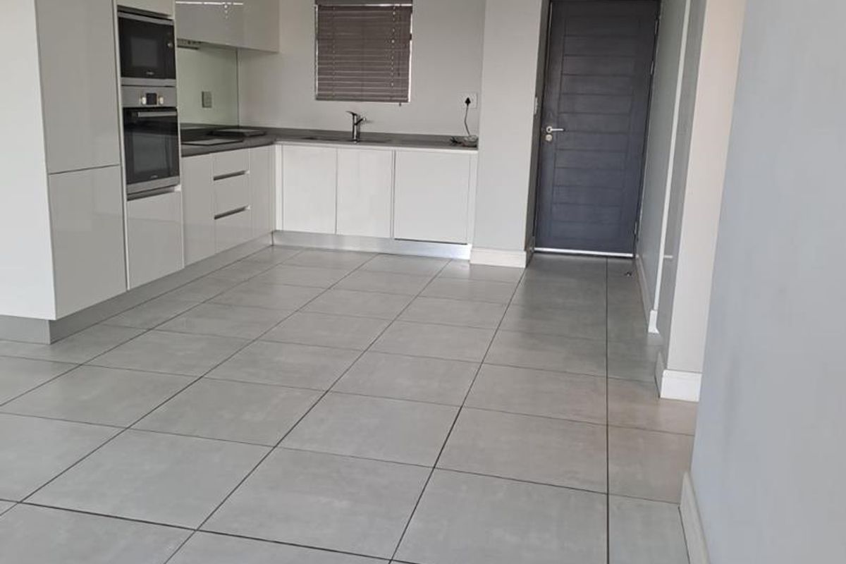 A modern, empty kitchen featuring white high-gloss cabinetry, built-in appliances, grey tiled flooring, and neutral walls, offering a clean and functional space.