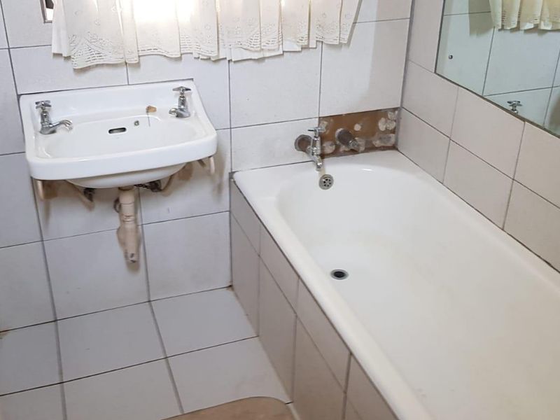 A compact bathroom featuring a wall-mounted basin and a standard bathtub, currently requiring cosmetic updates to the tiled wall area.