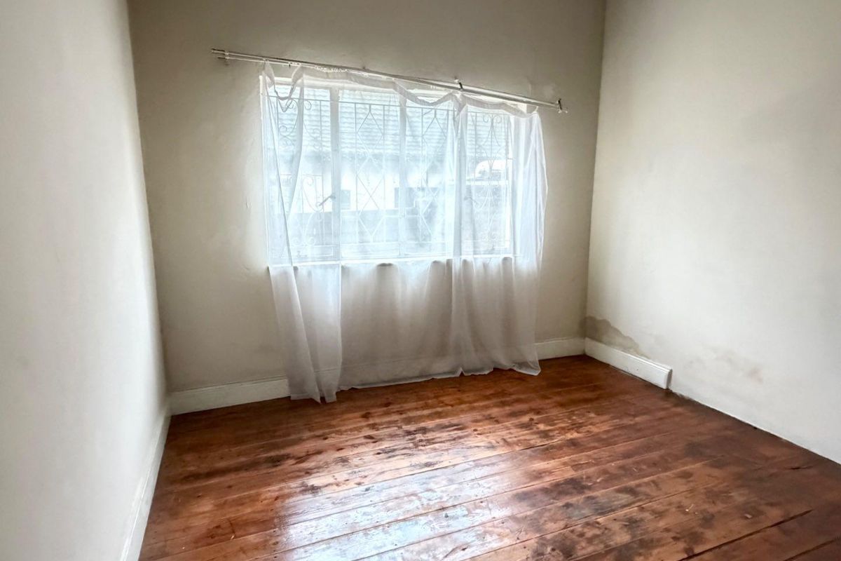 An empty bedroom featuring original timber flooring, a window with security bars, and visible signs of wall moisture damage.
