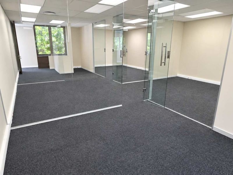 Modern open-plan office space featuring glass partitioning, suspended ceilings with recessed lighting, and clean carpeted flooring.