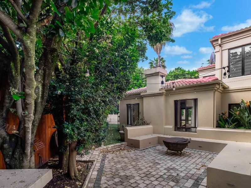 A private paved patio area featuring built-in seating, a chimney, and overhead string lighting, set against a lush garden backdrop.
