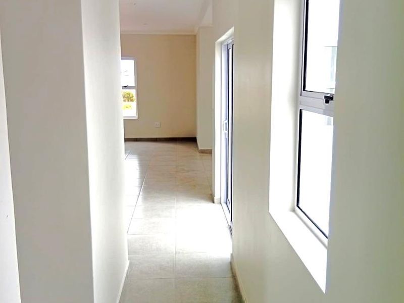 A bright, empty hallway featuring neutral tiled flooring, clean white walls, and natural light provided by aluminium-framed windows and a sliding door.