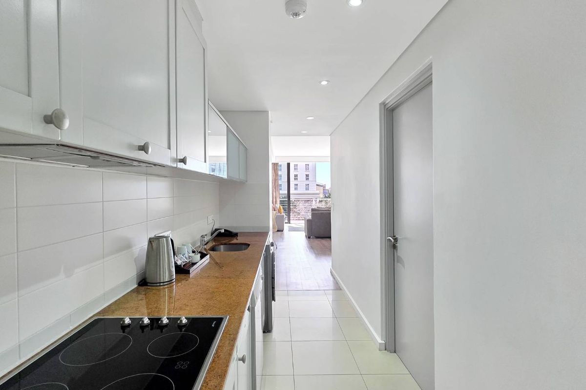 A modern galley-style kitchen featuring white cabinetry, stone countertops, an electric cooktop, and tiled flooring, leading towards an open-plan living area.