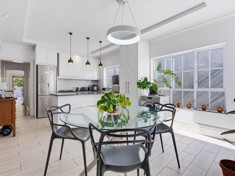 Modern open-plan kitchen and dining area featuring sleek white cabinetry, stone countertops, integrated appliances, and contemporary pendant lighting with bright, neutral finishes.