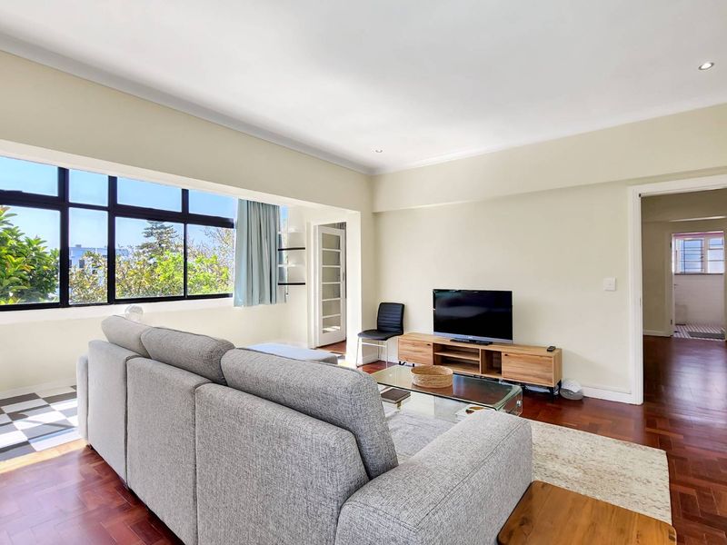 A bright and spacious living room featuring warm parquet flooring, large windows providing natural light, and a clean, neutral aesthetic.