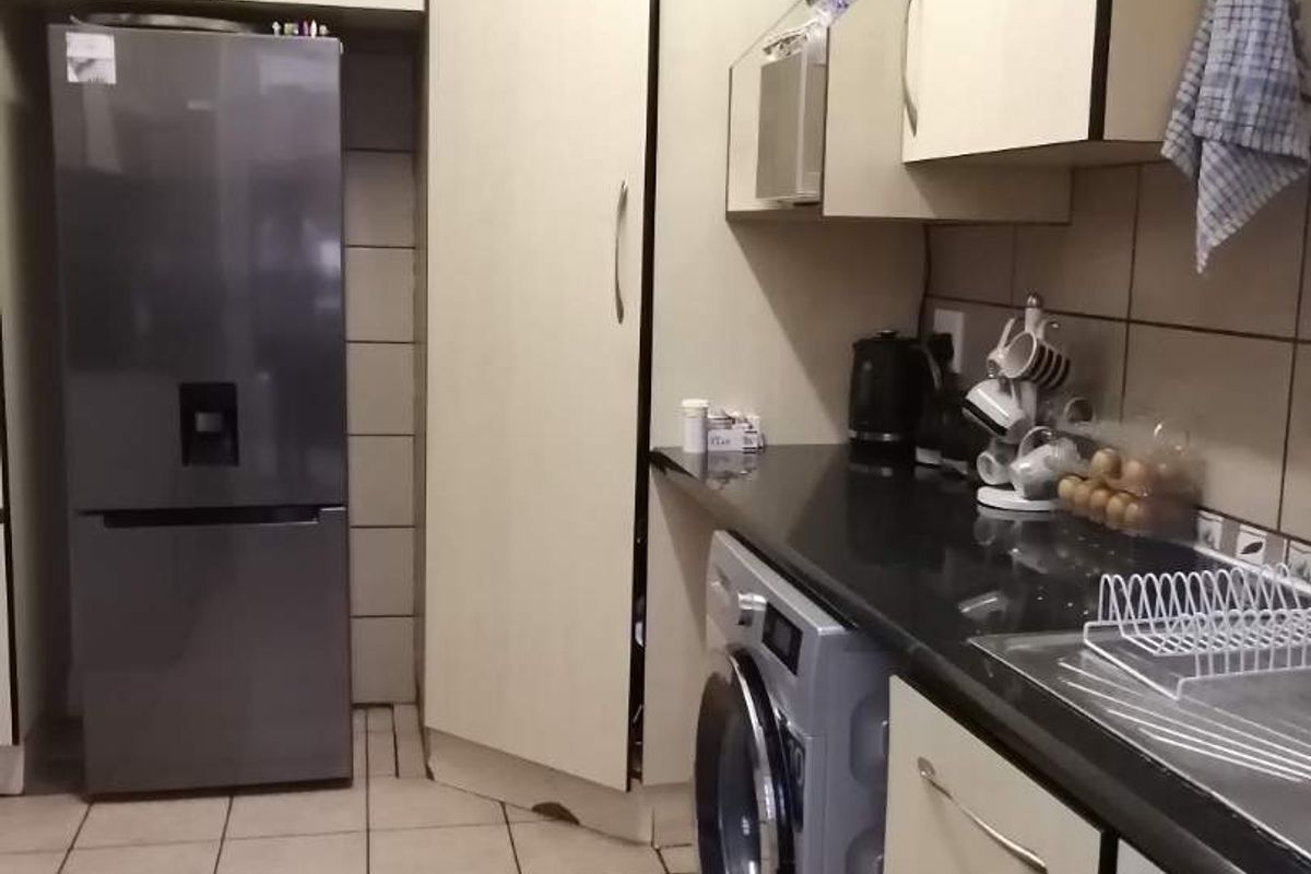 A compact kitchen featuring built-in cabinetry, dark countertops, tiled flooring, and integrated laundry plumbing, showing signs of wear and dated finishes.