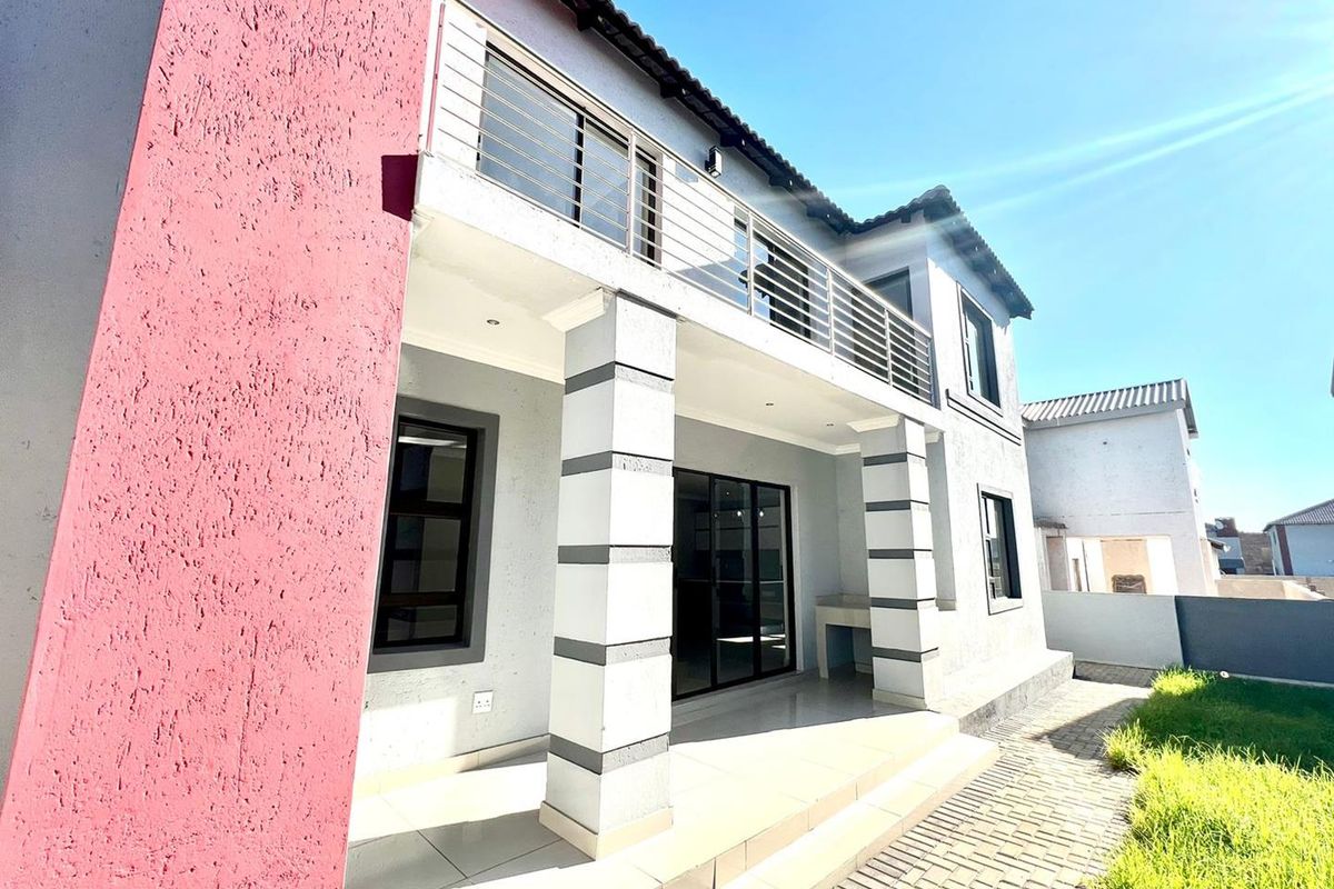 A modern double-story exterior featuring a covered patio, tiled flooring, a balcony with stainless steel railings, and a built-in outdoor braai area.