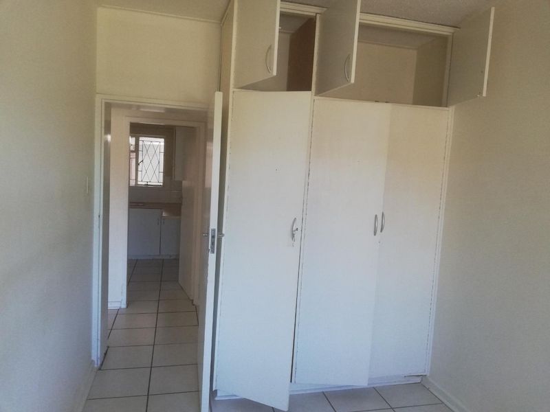 An empty bedroom featuring built-in cupboards and tiled flooring, with a view through to the kitchen area.