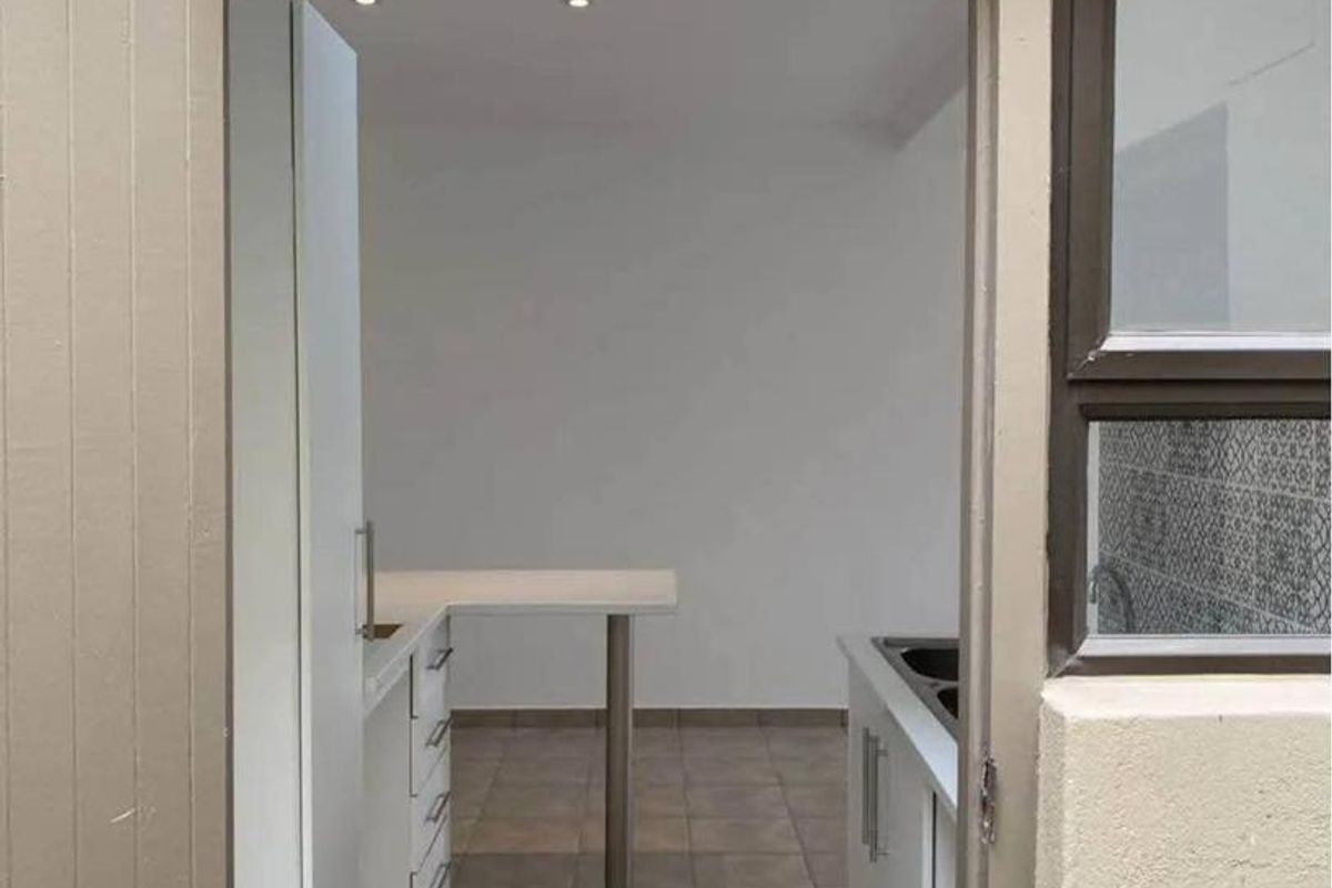 A view into a clean, empty kitchen featuring white built-in cabinetry, tiled flooring, and recessed lighting, accessed through an exterior doorway.