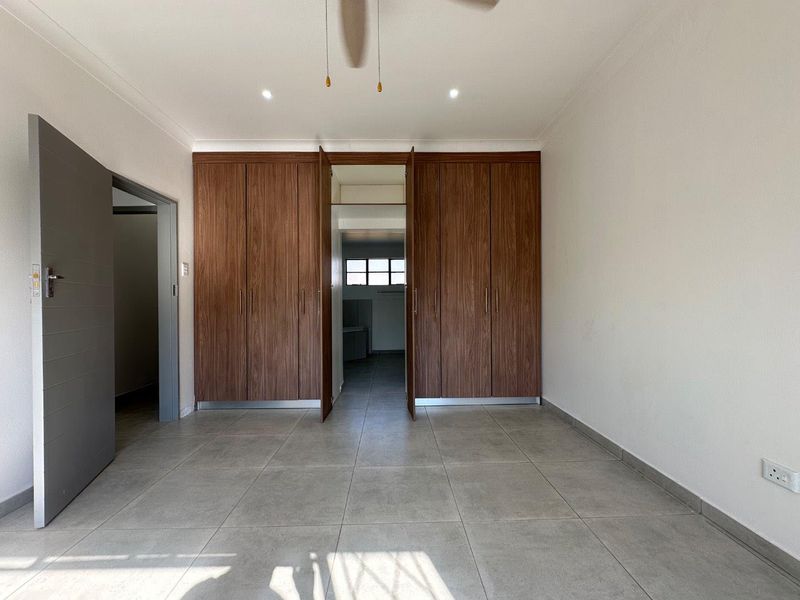A spacious, empty bedroom featuring modern tiled flooring, built-in wooden cupboards, and a ceiling fan, offering a clean and neutral living space.