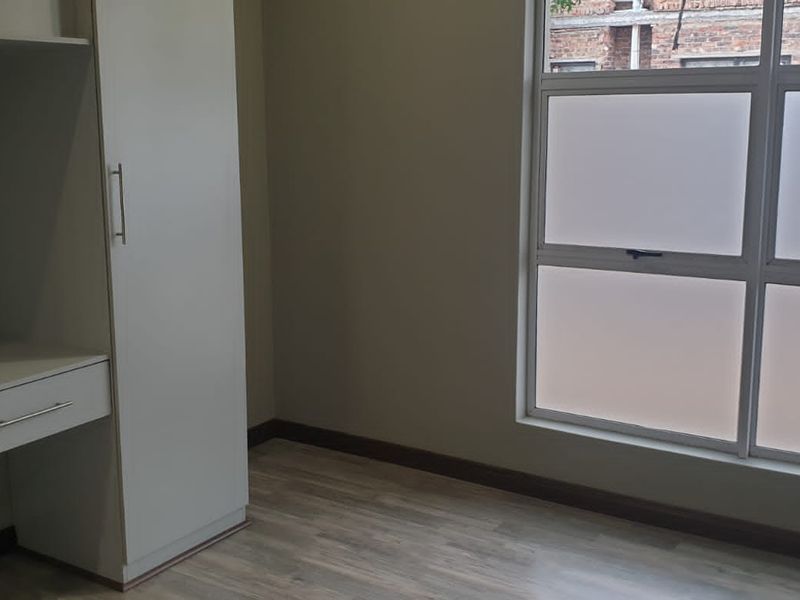 A bright, empty bedroom featuring modern laminate flooring, a built-in wardrobe, and a built-in desk with a large frosted glass window.