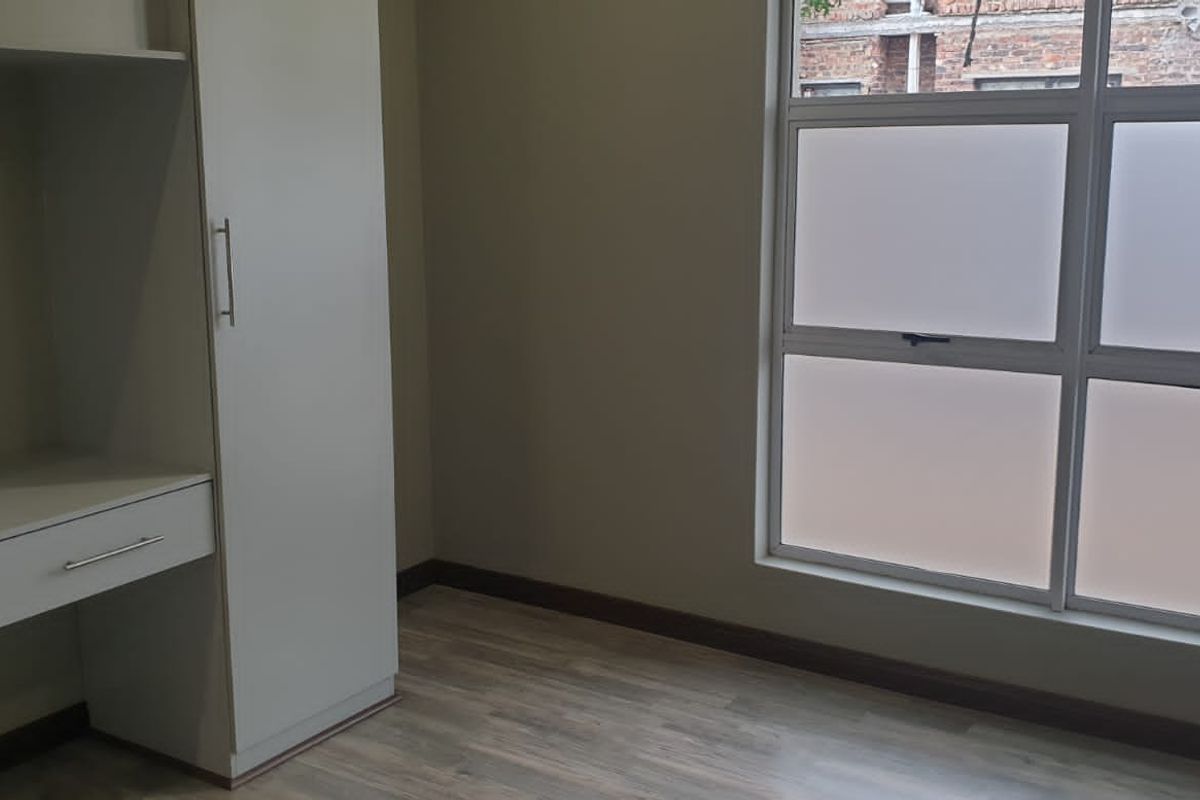 A bright, empty bedroom featuring modern laminate flooring, a built-in wardrobe, and a built-in desk with a large frosted glass window.