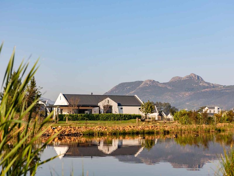 A scenic view of a modern residential property set against a backdrop of mountains, featuring a tranquil pond and well-maintained garden.