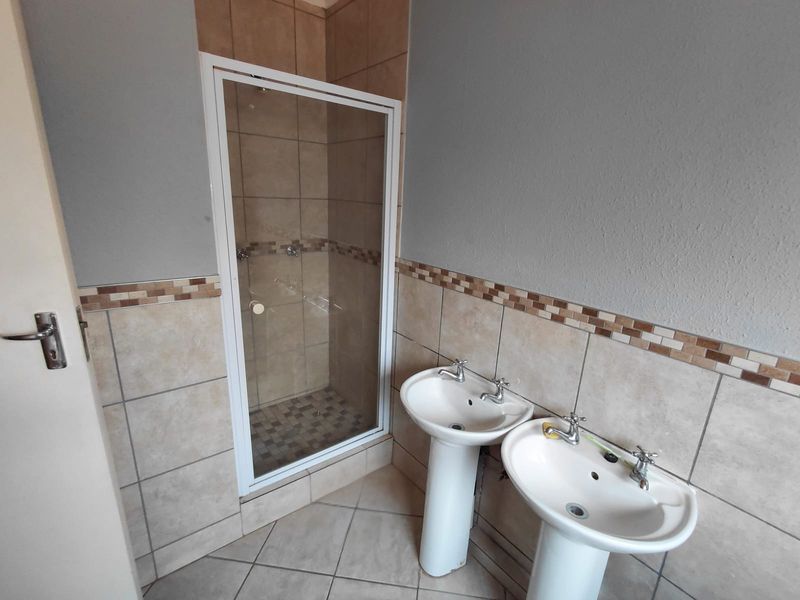 A functional bathroom featuring a shower enclosure and dual pedestal sinks with neutral wall and floor tiling.