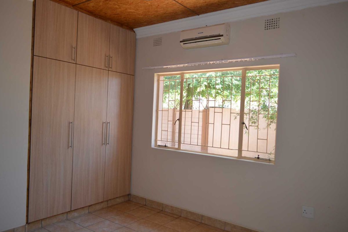 A bedroom featuring built-in wooden cabinetry, tiled flooring, a wall-mounted air conditioning unit, and a window with security bars.