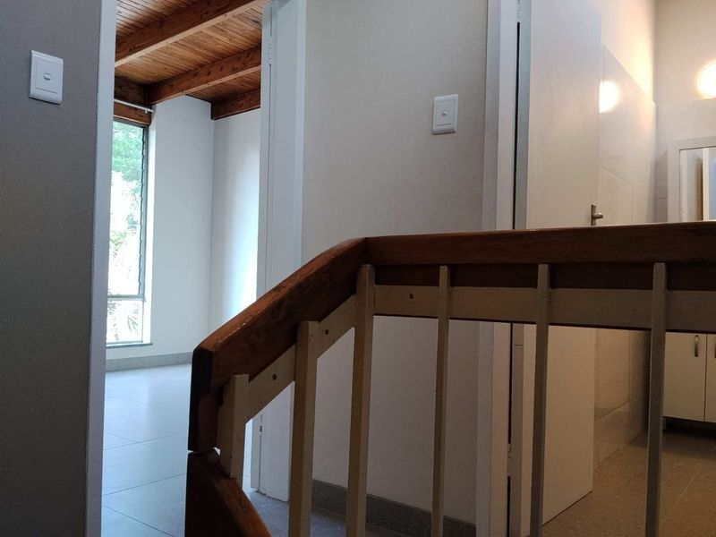 A bright hallway featuring a wooden staircase railing, exposed timber ceiling beams, and neutral tiled flooring leading to adjacent rooms.