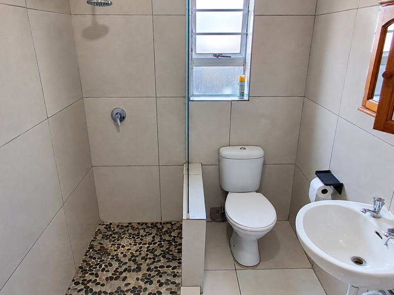 A compact, modern bathroom featuring a walk-in shower with a pebble-tiled floor, glass screen, pedestal basin, and neutral wall tiling.