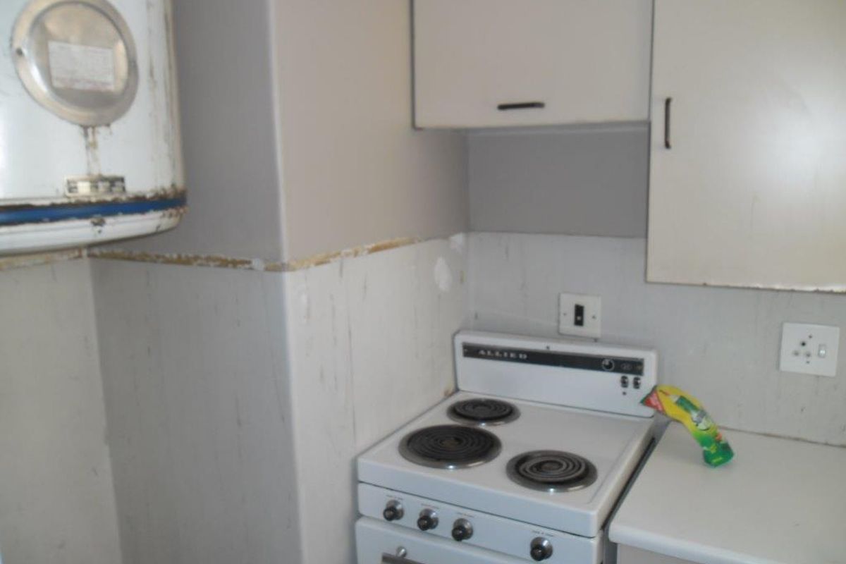 A compact kitchen area featuring an electric stove, wall-mounted water heater, and basic cabinetry, currently showing significant wear and requiring cosmetic updates.